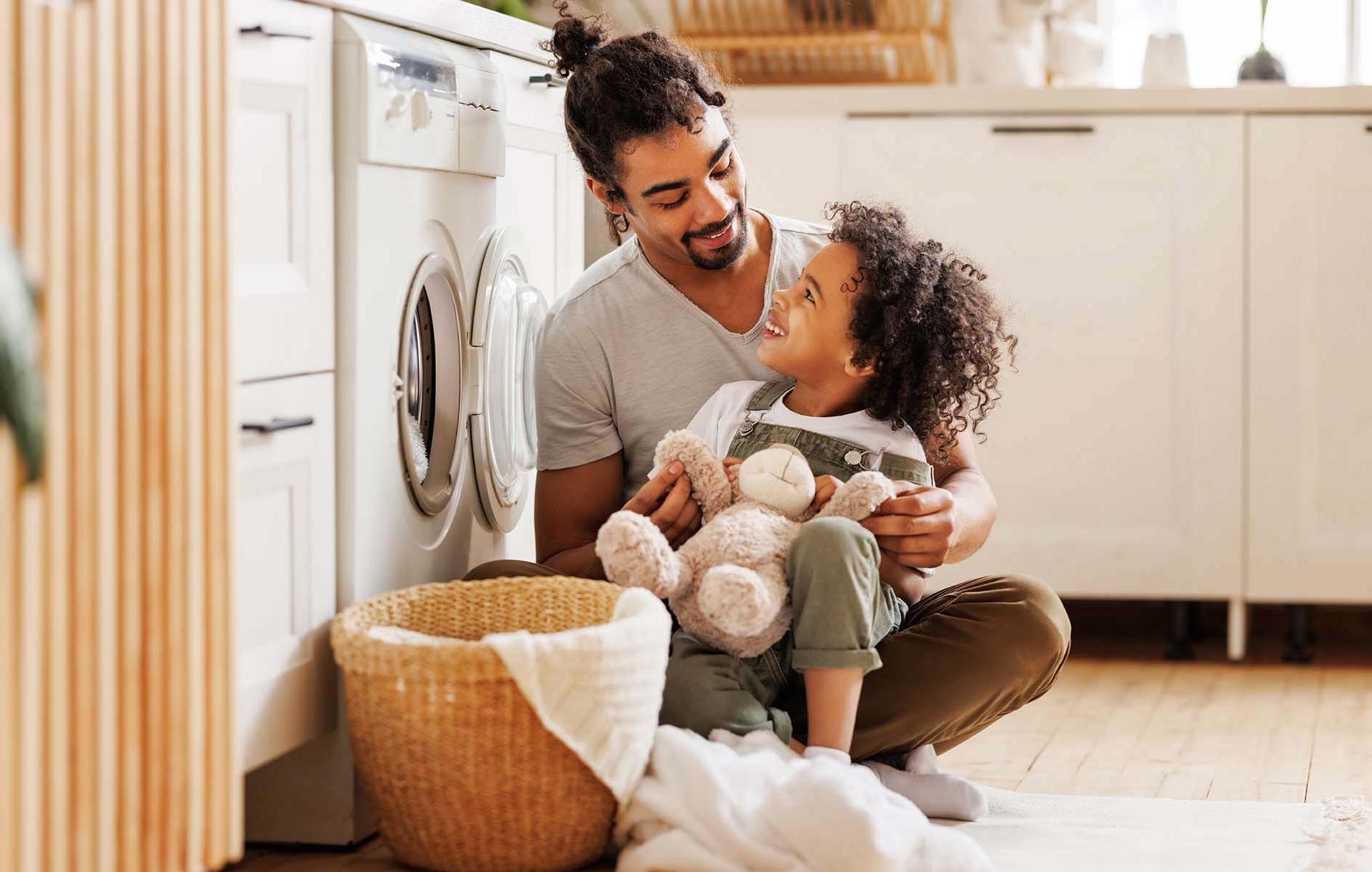 A man and a young child with a teddy bear smile at each other, sitting by a washing machine